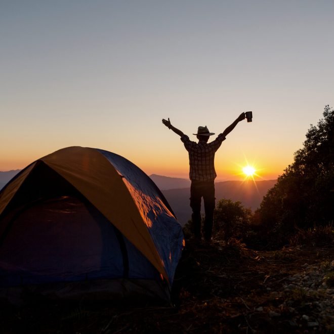 Silhouette of Happy man with holding coffee cup stay near tent around mountains under sunset light sky enjoying the leisure and freedom.